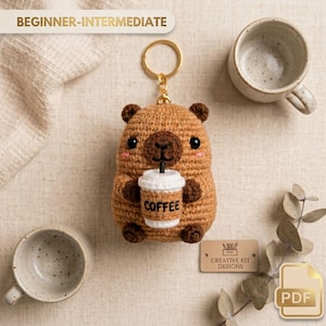Op de afbeelding: Een gehaakte bruine beren sleutelhanger met een koffiekopje met het woord "COFFEE" erop. De sleutelhanger heeft een gouden ring en is gelabeld "BEGINNER-INTERMEDIATE". Twee keramische mokken staan aan weerszijden van de beer.