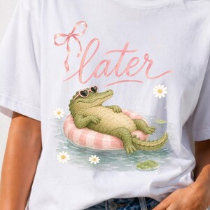May include: A white short-sleeve t-shirt featuring a graphic of a green alligator wearing heart-shaped sunglasses while floating on a pink and white striped inner tube in water, with the word later in pink script and a pink bow above it.