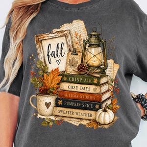 May include: A charcoal grey t-shirt featuring a graphic print of a stack of books labelled Crisp Air, Cozy Days, Autumn Stories, Pumpkin Spice, and Sweater Weather, topped with a lantern, a framed sign reading fall, and a white mug with a heart.