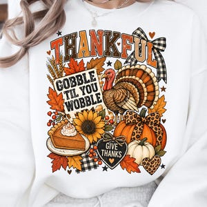 May include: A white long-sleeve sweatshirt featuring a graphic print with the text THANKFUL and GOBBLE TIL YOU WOBBLE. The design includes a turkey, a slice of pumpkin pie, a sunflower, pumpkins, autumn leaves, and a heart with GIVE THANKS.