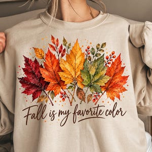 May include: A beige crewneck sweatshirt featuring a watercolor-style graphic of autumn leaves in shades of red, orange, yellow, and green, accented with small red berries. The text "Fall is my favorite color" is printed in a dark brown script font.
