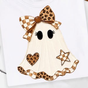 May include: A white t-shirt featuring a graphic of a ghost with black oval eyes and eyelashes. The ghost wears a large brown leopard print bow, a brown leopard print heart patch, a brown checkered star patch, and a brown checkered bottom hem.