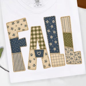 May include: A white t-shirt featuring the word FALL in large, block-style letters. Each letter is composed of various fabric patches in shades of navy blue, olive green, and cream, decorated with floral, plaid, and striped patterns with stitching.
