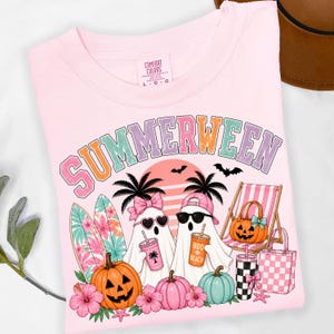 May include: A light pink t-shirt featuring the text SUMMERWEEN in a multi-colored font. The graphic includes two ghosts, one wearing a pink bow and heart sunglasses, the other a pink cap and black sunglasses, holding drinks. Also features surfboards, pumpkins, a beach chair, and a tote bag.