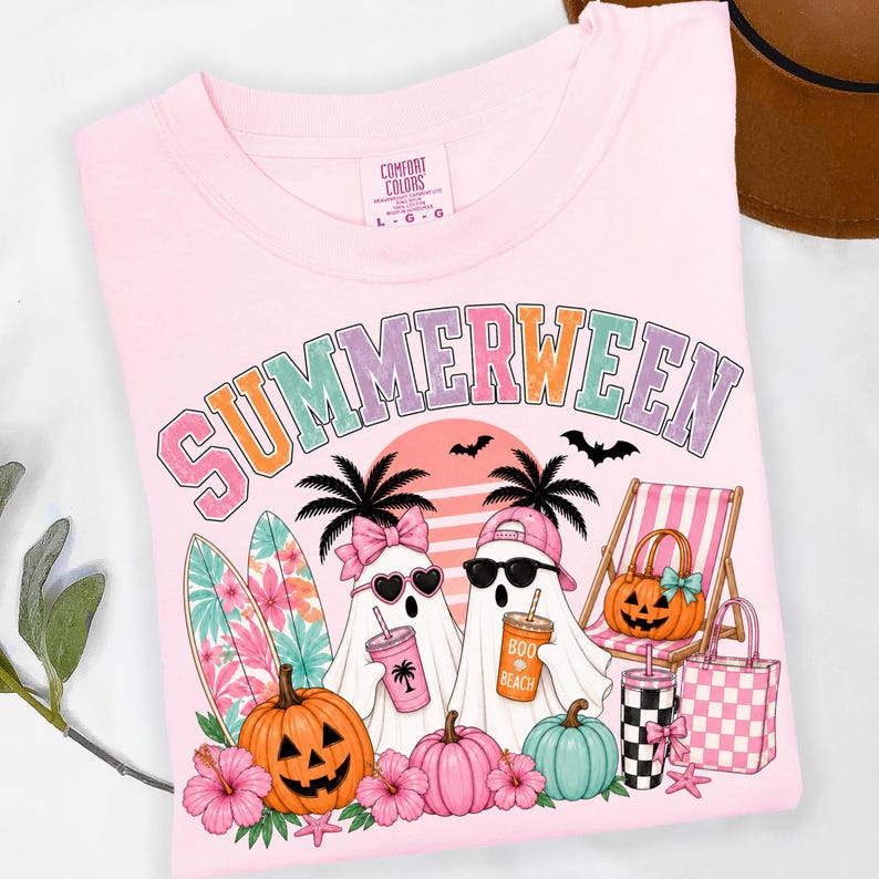 May include: A light pink t-shirt featuring the text SUMMERWEEN in a multi-colored font. The graphic includes two ghosts, one wearing a pink bow and heart sunglasses, the other a pink cap and black sunglasses, holding drinks. Also features surfboards, pumpkins, a beach chair, and a tote bag.