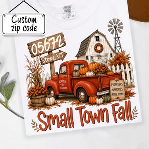 May include: A white t-shirt featuring a graphic of a red pickup truck loaded with pumpkins, a barn, a windmill, and a wooden sign reading 05672 Stowe, VT. The text Small Town Fall appears at the bottom, with a sign listing Pumpkins, Hayrides, Apple Cider.