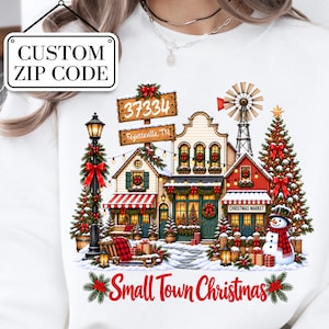 Könnte beinhalten: Ein weißes Sweatshirt mit einer Grafik einer festlichen Stadtszene mit Schneemann, Weihnachtsbaum, Windmühle und weihnachtlich geschmückten Gebäuden. Der Text lautet 37334, Fayetteville, TN, und Small Town Christmas in roter Schrift.