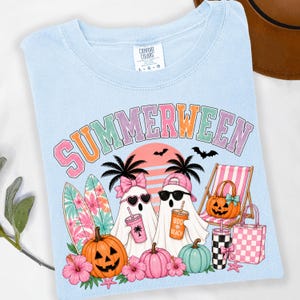 May include: A light blue t-shirt featuring a graphic with the text SUMMERWEEN in pastel colors. The design includes two ghosts, surfboards, palm trees, a beach chair, pumpkins, a tote bag, and a drink cup labeled BOO BEACH.