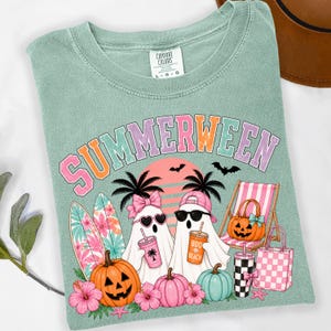 May include: A sage green t-shirt featuring the text SUMMERWEEN in colorful letters. The graphic includes two ghosts, one wearing a pink bow and heart sunglasses, the other a pink cap and black sunglasses, holding drinks, with surfboards, pumpkins, and a beach chair.