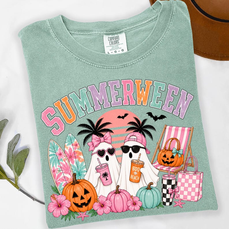 May include: A sage green t-shirt featuring the text SUMMERWEEN in colorful letters. The graphic includes two ghosts, one wearing a pink bow and heart sunglasses, the other a pink cap and black sunglasses, holding drinks, with surfboards, pumpkins, and a beach chair.