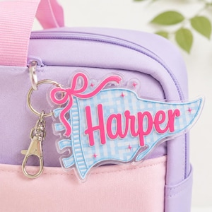 May include: A clear acrylic pennant-shaped keychain featuring the name Harper in bright pink script, accented with a pink bow and a light blue gingham pattern. The keychain is attached to a silver metal split ring and a lobster clasp.