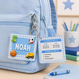May include: A light blue backpack with a square, light blue gingham-patterned luggage tag attached by a silver metal clip. The tag features the name NOAH in white letters, with small dinosaur, rocket, basketball, and truck icons attached.
