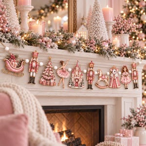 Op de afbeelding: Een decoratieve slinger met een reeks hangende ornamenten, waaronder een roze slee, notenkrakers, een kerstboom, een ballerina, een kasteel, een hobbelpaard, een muis in een japon en een meisje, verbonden door een snoer van witte kralen.