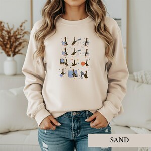 May include: A sand-colored crewneck sweatshirt featuring a graphic print of multiple black ducks engaged in various astronomical activities, including using telescopes, observing planets, and reading books, scattered with small star icons.