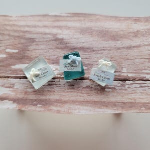 Pode incluir: Três sabonetes pequenos com etiquetas. Um é um cubo transparente com um laço branco e uma etiqueta que diz "Soft Clean Soap". Outro é um sabonete verde-azulado com um laço branco e uma etiqueta que diz "Sea Glass Soap". O terceiro é um sabonete transparente com um laço branco e uma etiqueta que diz "Fresh Linen Soap".