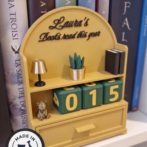 Può includere: Una libreria giallo senape con la scritta "Laura's Books read this year". Presenta una lampada in miniatura, una piccola statuina di orso, una pianta in vaso e blocchi numerati che mostrano "015". Un cassetto è in basso. La libreria è ambientata su uno sfondo di libri.