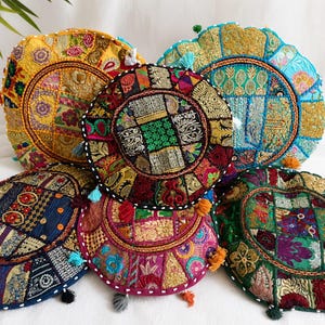 May include: A collection of five round floor cushions featuring intricate patchwork designs with vibrant embroidery, metallic threads, and small tassels. Each cushion displays a unique combination of floral and geometric patterns in yellow, blue, green, red, and gold.