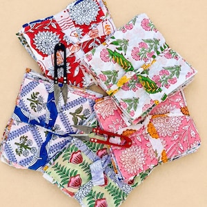 May include: A collection of folded fabric squares featuring various floral and botanical block print patterns in red, blue, pink, green, and yellow. Two pairs of metal thread snips with black and red handles are placed on top of the fabric stacks.