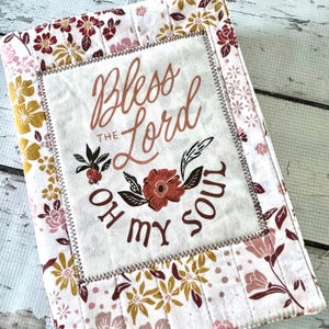 May include: A quilted fabric book cover featuring a floral pattern in shades of pink, mustard yellow, and maroon. A central rectangular patch displays the text Bless The Lord O My Soul in brown script and block lettering with a floral graphic.