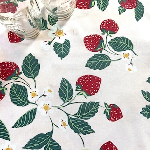 May include: A white tablecloth with a repeating pattern of red strawberries, green leaves, and white flowers. Two clear glass jars are on the tablecloth.
