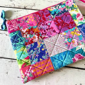 May include: A quilted zipper pouch constructed from a patchwork of triangular fabric pieces in vibrant floral and geometric patterns. The bag features a bright pink zipper closure and a small teal fabric tie attached to the side zipper pull.