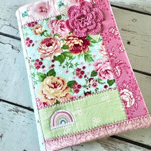 May include: A rectangular fabric book cover featuring a patchwork design of floral patterns in shades of pink, green, and light blue. The cover is embellished with a large, dark pink crocheted flower and a small, embroidered rainbow patch.