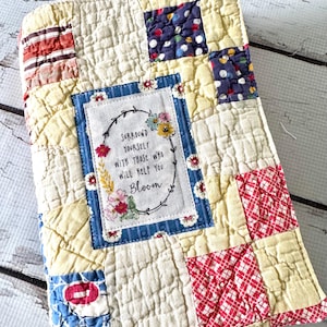 May include: A quilted book cover with a patchwork design. The cover features squares of various fabrics in cream, red, blue, and patterned prints. A central patch displays the text "Surround yourself with those who will help you Bloom" within a floral wreath.