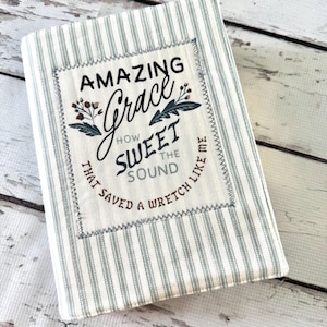 May include: A rectangular fabric cover featuring a light cream and grey striped pattern. A central rectangular patch is stitched on with the text Amazing Grace How Sweet The Sound That Saved A Wretch Like Me and floral accents.