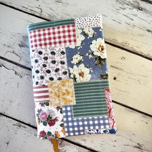 May include: A patchwork fabric book cover with a variety of patterns, including gingham, floral, and striped designs. The cover features a yellow bookmark and is made of various fabric squares sewn together. The color palette includes red, blue, green, and yellow.