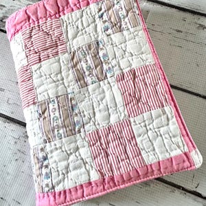 May include: A rectangular quilted fabric cover featuring a patchwork design of alternating white, red-striped, and floral-patterned squares. The entire piece is bordered by a solid pink fabric trim and finished with visible white stitching throughout.