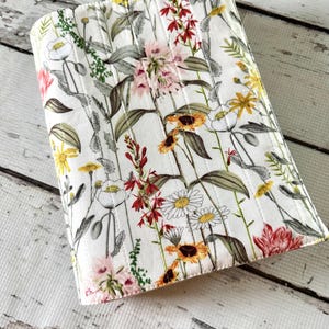 May include: A rectangular fabric book cover featuring a white background with a colorful botanical print of various wildflowers including daisies, sunflowers, and red blossoms. The cover is quilted with vertical stitching lines throughout.