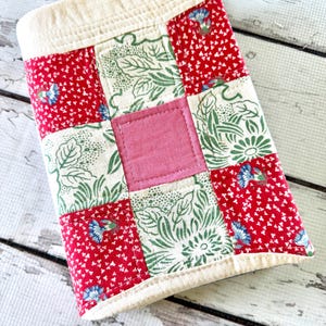 May include: A rectangular fabric cover featuring a patchwork design of red floral squares, green leaf-patterned squares, and a solid pink center square. The piece is finished with a cream-colored fabric border and visible quilted stitching throughout.