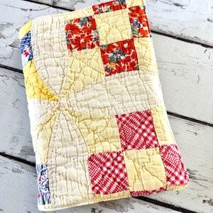 May include: A handmade, quilted notebook cover with a patchwork design. The cover features squares of red floral fabric, red and white checkered fabric, and yellow fabric. The cover is made of a soft, quilted material.
