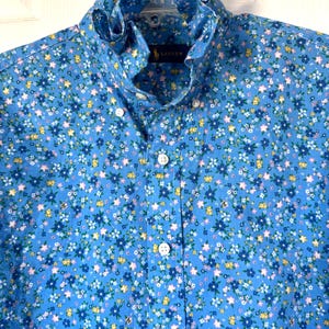 May include: A blue long-sleeve button-down shirt featuring a small-scale floral pattern in white, yellow, and dark blue. The shirt has a high, ruffled collar and a front button placket with white buttons. A Lauren label is visible at the neck.