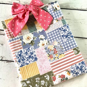 May include: A patchwork fabric notebook cover with a pink polka dot bow. The cover features various floral and patterned fabric squares in shades of blue, pink, green, and yellow. The bow is tied at the top.