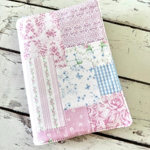 May include: A patchwork journal cover featuring a variety of floral and patterned fabrics in shades of pink, white, and blue. The cover is rectangular with rounded corners, and the fabrics are sewn together in a patchwork design.