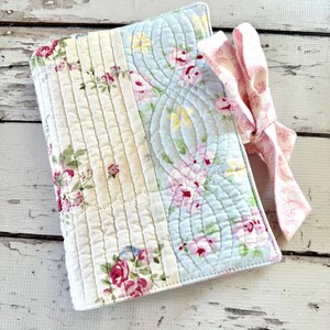 May include: A rectangular quilted fabric book cover featuring a patchwork design of cream and light blue panels with floral patterns in pink, green, and yellow. The cover includes a pink and white patterned fabric ribbon tied in a bow on the side.