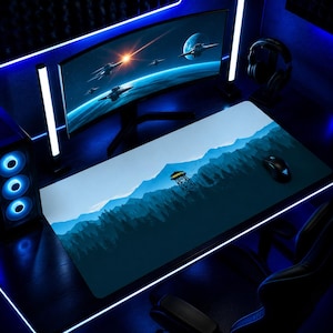 Puede incluir: Una alfombrilla de ratón rectangular grande con un diseño de paisaje de montaña azul. La alfombrilla presenta una ilustración detallada de un bosque, montañas y una torre de vigilancia. Un monitor de ordenador, un ratón y unos auriculares son visibles en el fondo.