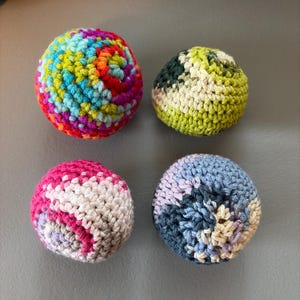May include: Four spherical objects made of crocheted yarn. One ball features a vibrant multicolored pattern of red, blue, yellow, and green. Another is a mix of light and dark green. The third is pink, white, and gray. The fourth is blue and white.