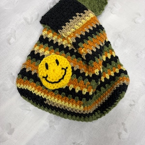 May include: A crochet bag featuring a multi-coloured striped pattern in black, olive green, mustard yellow, and cream. The bag has a prominent, circular, bright yellow smiley face patch with a black embroidered smile and eyes stitched on the front.