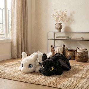 May include: Two plush dragon toys, one white and one black, are lying on a woven rug. The white dragon has large blue eyes, while the black dragon has yellow eyes. The toys are in a room with neutral tones and a window.