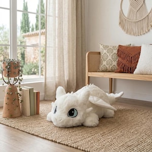 May include: A large, white plush dragon toy with blue eyes and small wings rests on a woven jute rug. The dragon is in a relaxed pose, with its head and paws forward. The room has a neutral color palette with natural light.