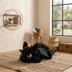 May include: A large, plush black dragon-shaped pillow with yellow eyes and black wings rests on a woven jute rug. The pillow is a soft, cuddly toy. The room has a neutral color palette with a wooden shelf and a small wooden stool.
