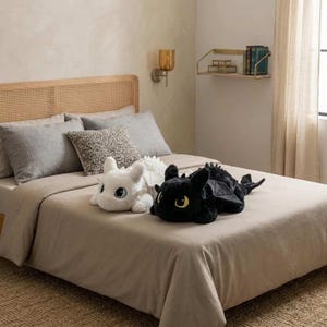 May include: Two plush dragon toys, one white and one black, rest on a bed with a tan comforter. The white dragon has large blue eyes, while the black dragon has yellow eyes. The bed is made up with gray pillows and a woven headboard.