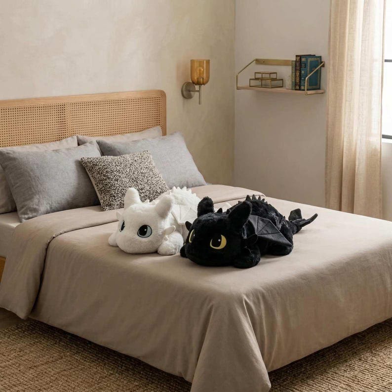 May include: Two plush dragon toys, one white and one black, rest on a bed with a tan comforter. The white dragon has large blue eyes, while the black dragon has yellow eyes. The bed is made up with gray pillows and a woven headboard.