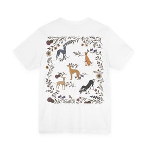 May include: A white short-sleeved t-shirt featuring a central rectangular graphic on the back. The design displays five grey, tan, and black greyhound dogs surrounded by a detailed botanical border of brown, purple, and green flowers.