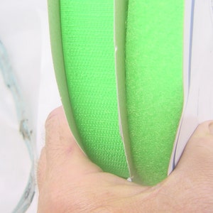 May include: Two rolls of bright green hook and loop fastener tape. The tape is folded in half and held in a hand.