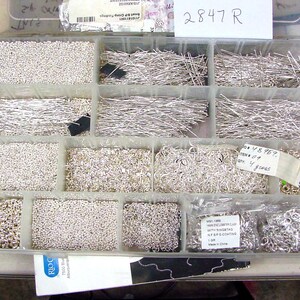 May include: A clear plastic storage box filled with various silver jewelry findings, including beads, jump rings, and head pins. The box is labeled "2847 R".