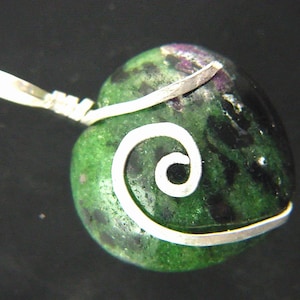 Op de afbeelding: Een hartvormige hanger in groen en zwart met een zilveren draad eromheen gewikkeld in een spiraalvorm. De hanger heeft een zilveren lus om aan een ketting te bevestigen.