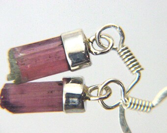 May include: A pair of silver earrings featuring pink tourmaline stones. The stones are set in silver bezels and hang from silver spring rings.